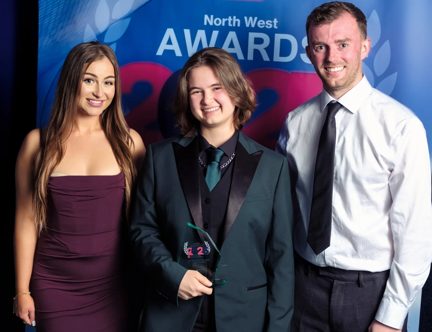 Alex centre of the picture in a dark green 3 piece suit and tie holding her award and smiling to camera.