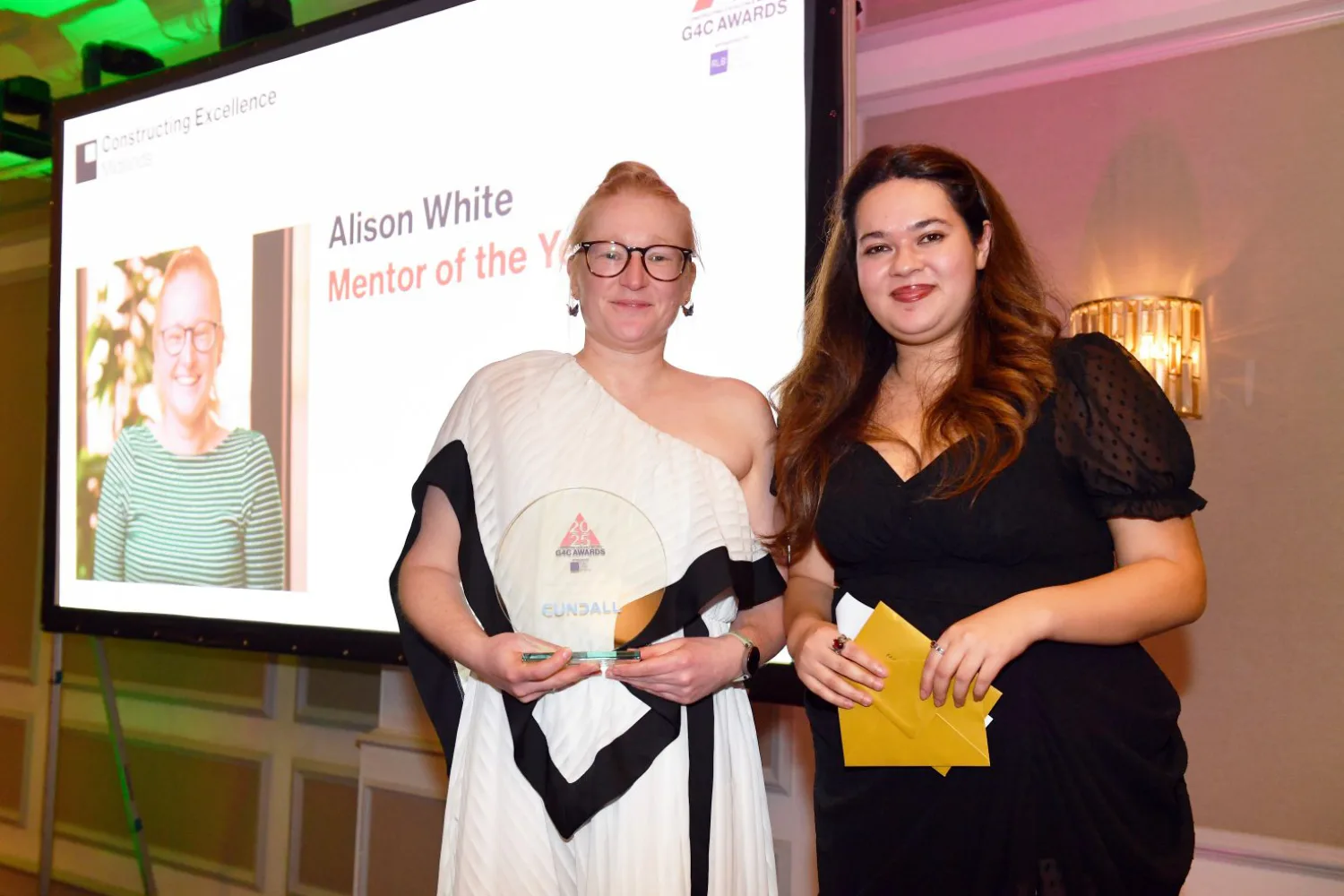 Alison on stage in a long white dress holding her award with the host in front of the projection screen