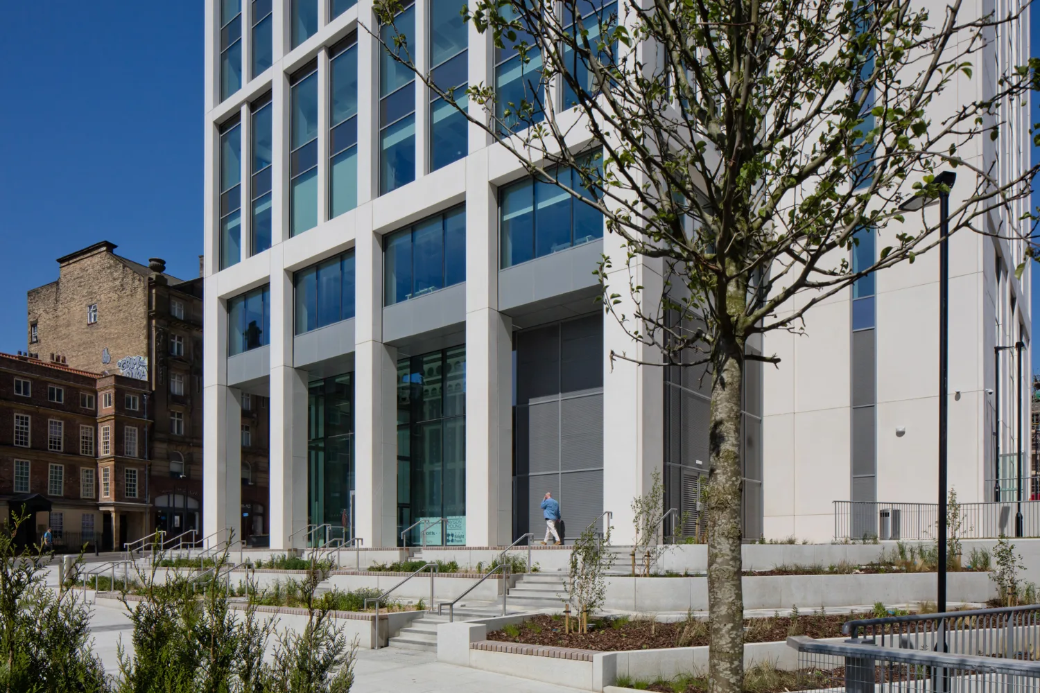 A modern multi‑storey glass and concrete building with large windows and a landscaped entrance featuring steps, railings, planted areas, and a tree in the foreground. Two people walk near the entrance.