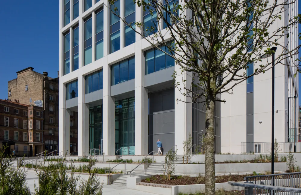 A modern multi‑storey glass and concrete building with large windows and a landscaped entrance featuring steps, railings, planted areas, and a tree in the foreground. Two people walk near the entrance.