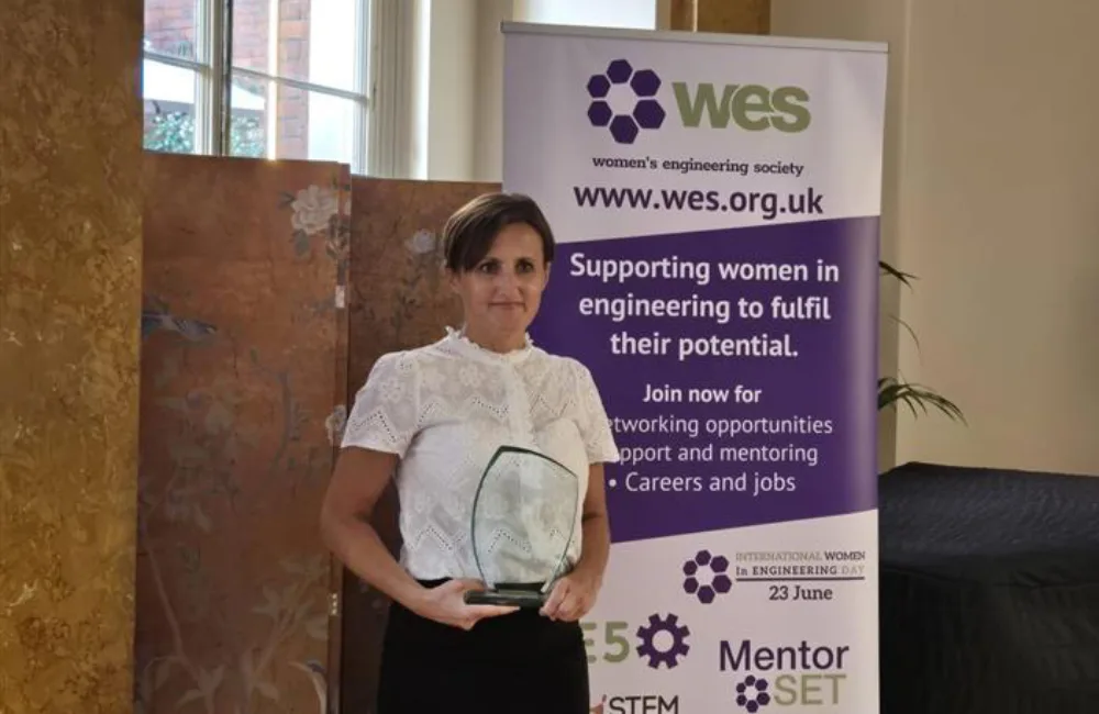 Person holding a glass award stands in front of a Women's Engineering Society banner promoting support, mentoring, and careers for women in engineering. Logos for WES, Mentor SET, and International Women in Engineering Day are visible.