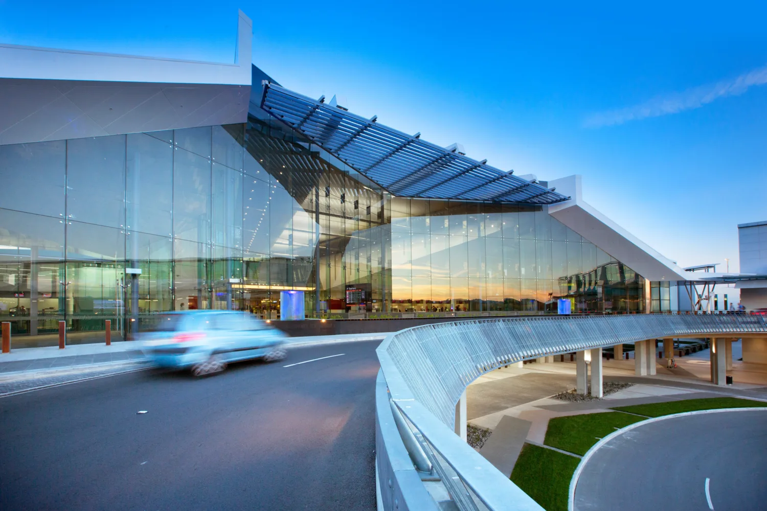 A modern airport building with large glass windows and a metal roof structure stands under a clear blue sky. A blue car drives on a curved road in front of the building.