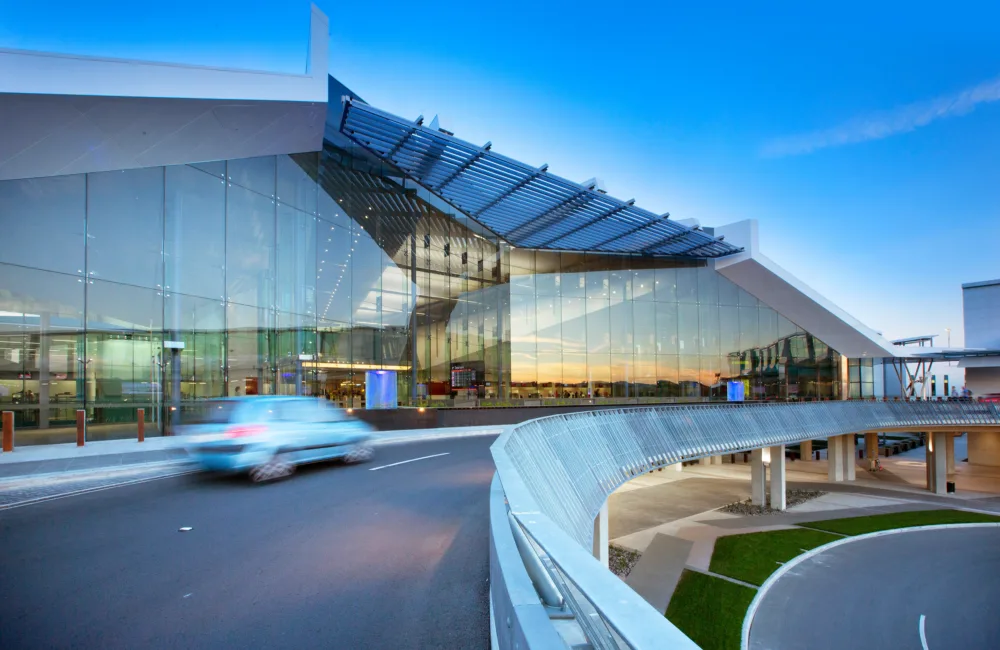 A modern airport building with large glass windows and a metal roof structure stands under a clear blue sky. A blue car drives on a curved road in front of the building.
