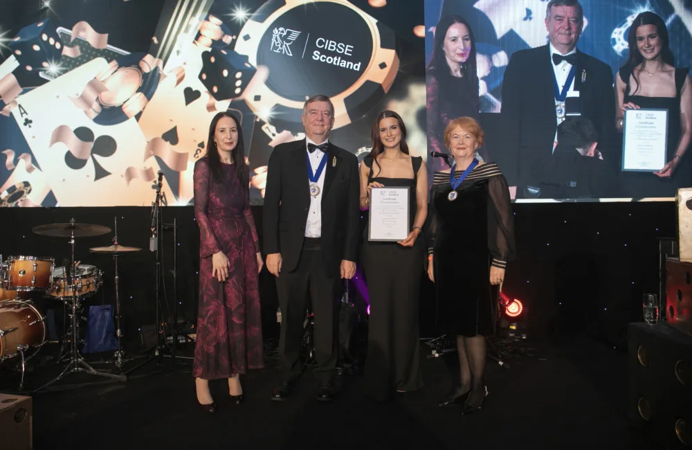 Four people standing on a stage in formal attire, two wearing medals. One person holds a framed certificate. Behind them is a large screen displaying ‘CIBSE Scotland’ with playing card graphics. A drum set is visible on the left.