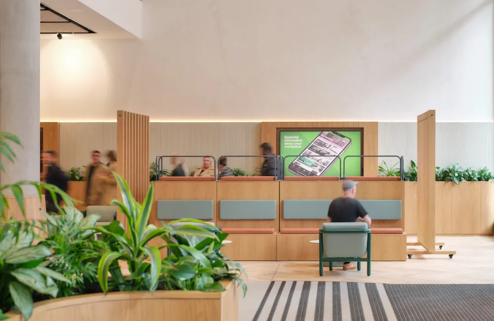 Interior of a modern lobby with light wood partitions, built-in bench seating, and potted plants. Several people walk past while one person sits on a chair. A wall display shows a smartphone graphic with text about workplace information.