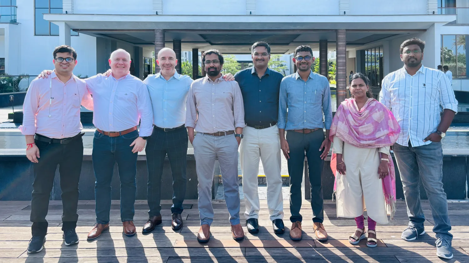 Employees stand in a row on a wooden deck outdoors with Chennai office visitors. A modern building is behind them.