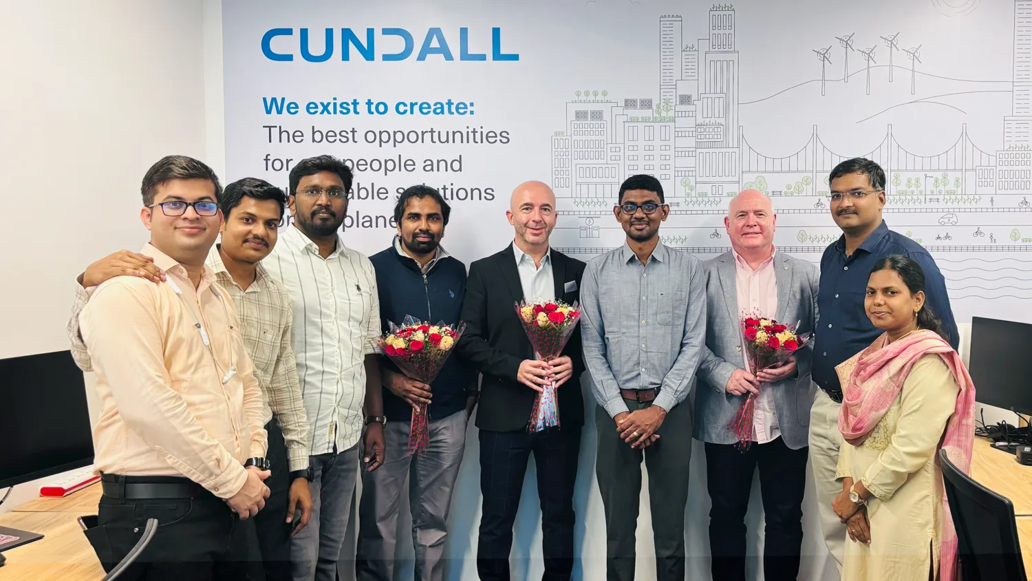 Employees stand in Chennai office, visitors are holding flower bouquets. Behind them, a wall displays the mission statement and line art illustration. Desks with monitors are visible on both sides.