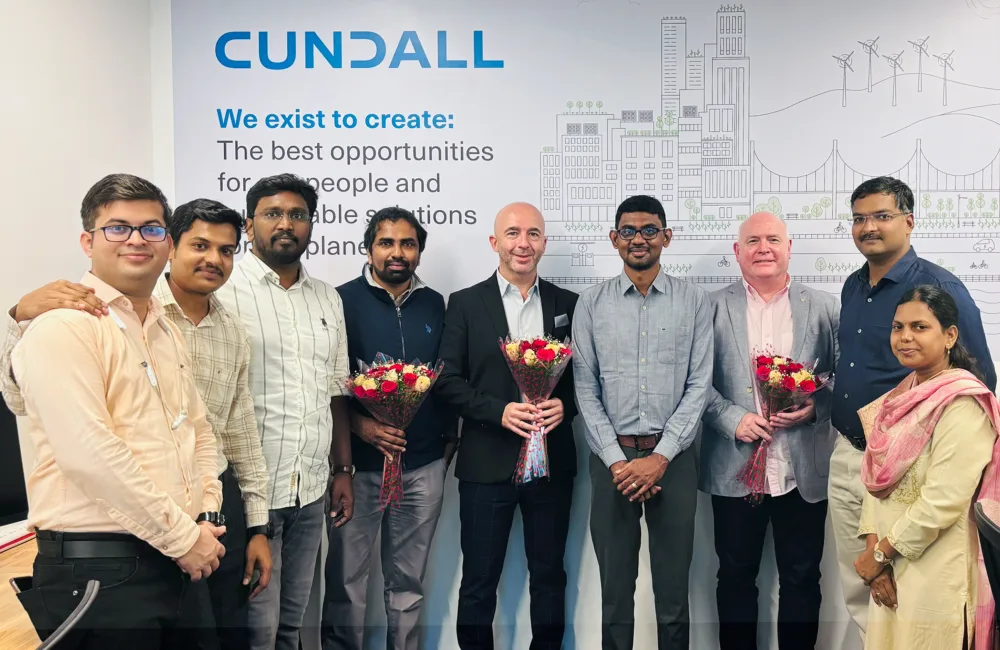 Employees stand in Chennai office, visitors are holding flower bouquets. Behind them, a wall displays the mission statement and line art illustration. Desks with monitors are visible on both sides.