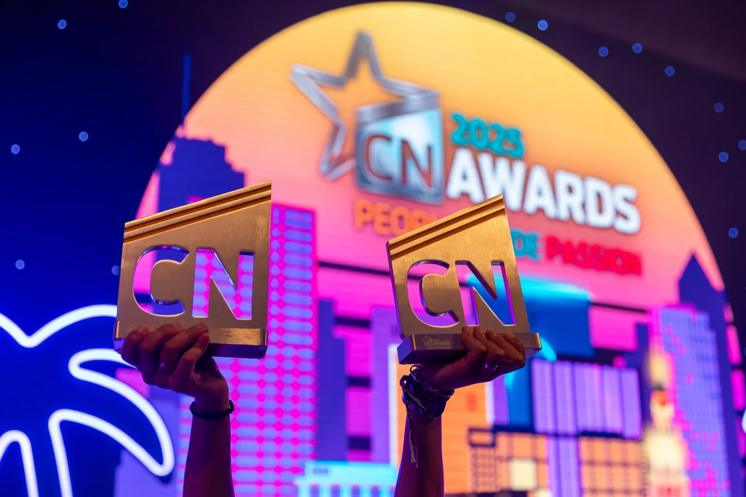 Two hands holding golden trophies engraved with 'CN' in front of a colourful backdrop showing a cityscape and the text '2023 CN AWARDS PEOPLE DE PASION' with a star logo.