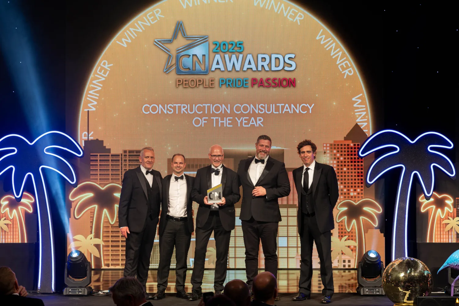 Five men in suits stand on a stage holding an award. Behind them is a circular sign with '2025 CN AWARDS,' 'PEOPLE PRIDE PASSION,' and 'CONSTRUCTION CONSULTANCY OF THE YEAR.' Neon palm trees flank the stage