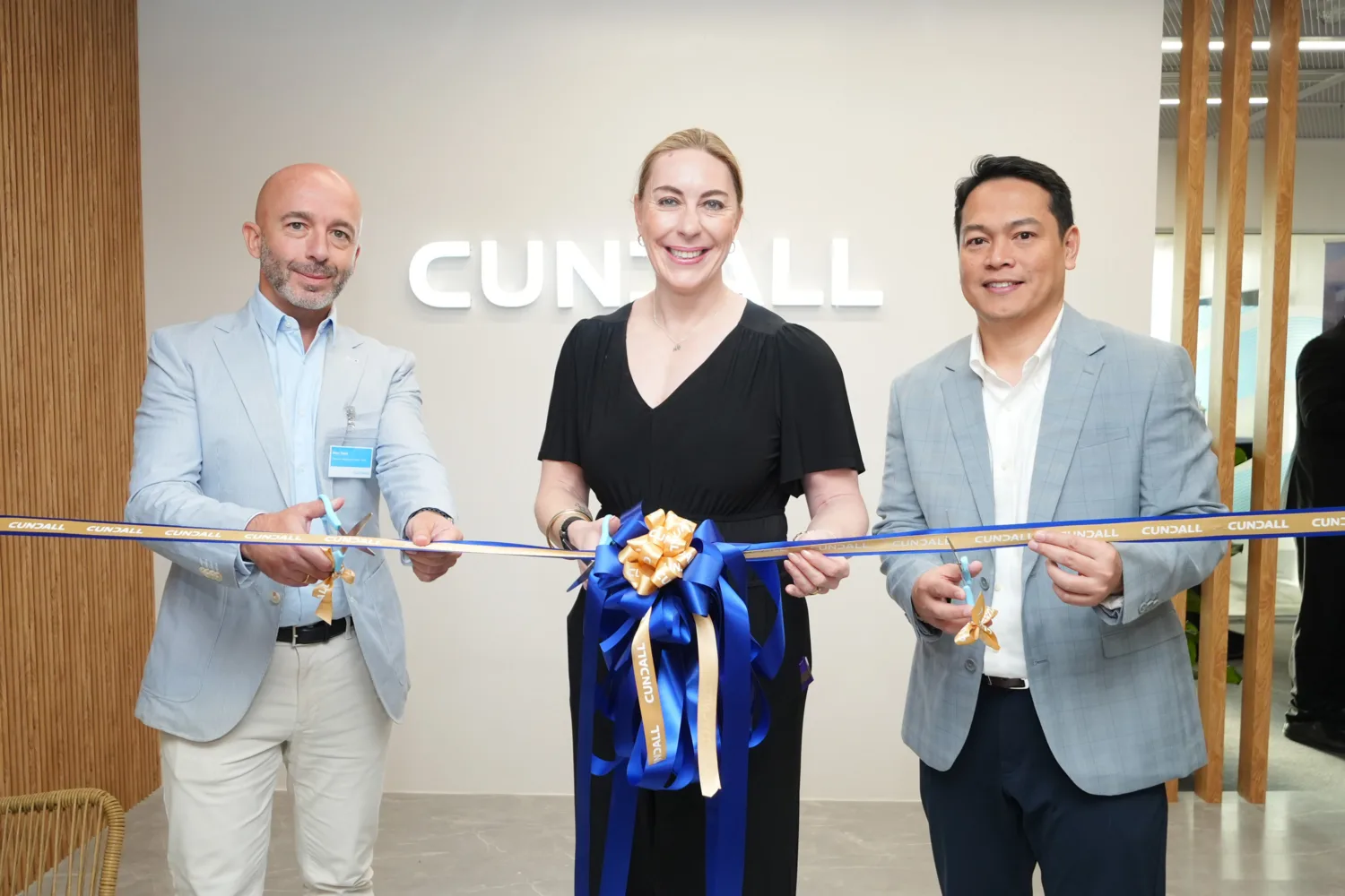 Three people stand cutting a ceremonial ribbon decorated with blue and gold bows. They are in front of a wall featuring the text “CUNDALL,” inside a modern office space.