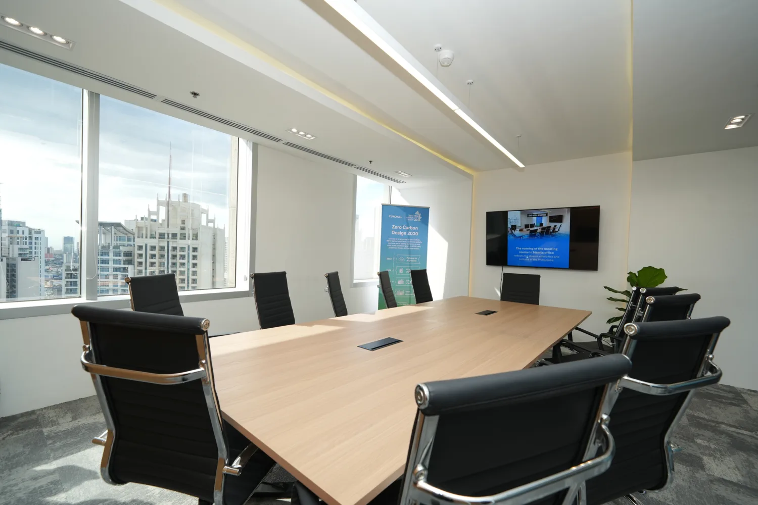 A modern conference room with a large wooden meeting table surrounded by black chairs. Floor‑to‑ceiling windows show a city skyline. A screen on the wall displays a presentation beside a standing banner.