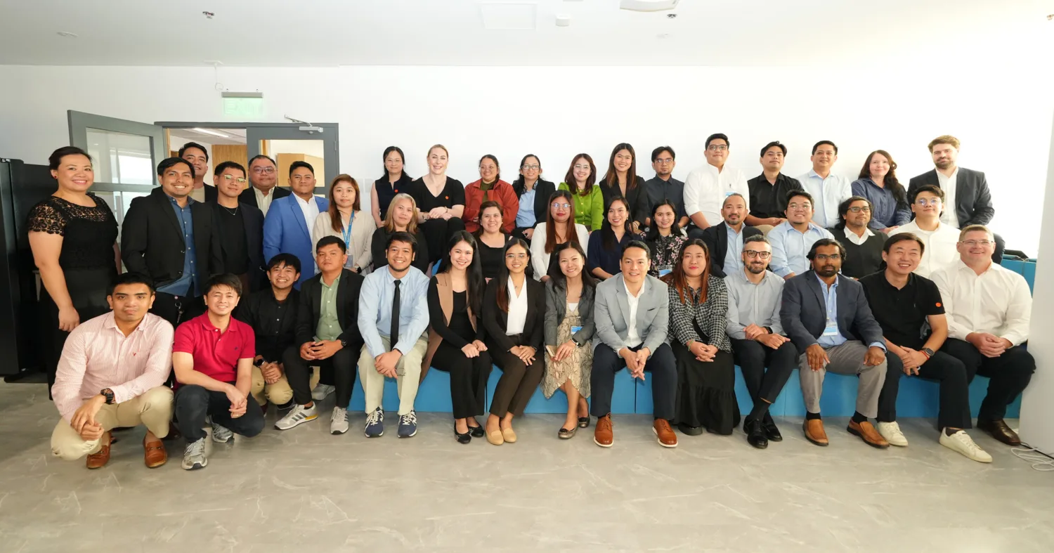 A large group of people pose together in a bright office space, arranged in three rows. They stand and sit in front of a white wall and blue seating, with a projector mounted on the ceiling above.