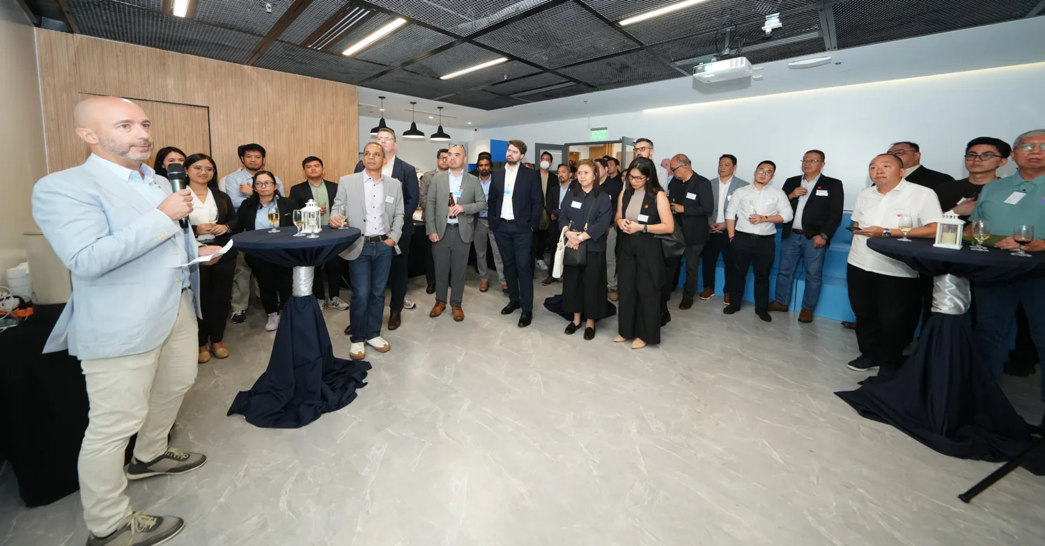 A large group of people stand in an office event space, listening to a speaker holding a microphone. High tables with drinks are scattered around the room, which features a patterned ceiling and bright lighting.