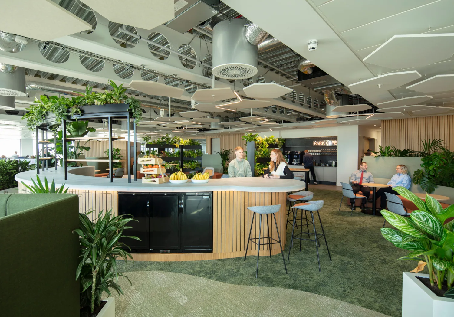 Modern office space with a curved white counter featuring stools, fruit bowls, and a display stand. Surrounding the counter are green plants, wooden slatted panels, and seating areas with tables and chairs. The ceiling has exposed ducts, circular vents, and suspended acoustic panels.