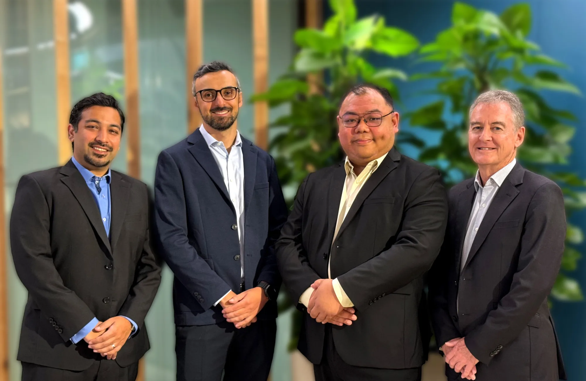 Four people stand indoors wearing dark business suits in front of tall green plants and wooden vertical panels. They are positioned in a row, facing forward in a modern office setting.