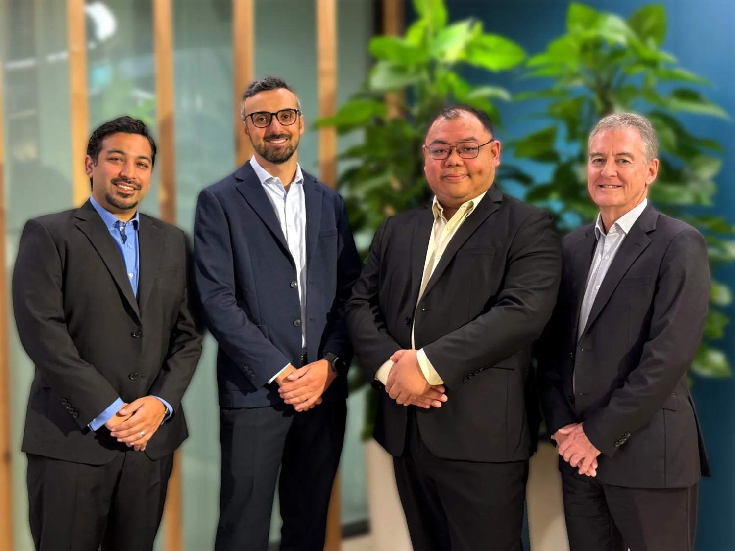 Four people stand indoors wearing dark business suits in front of tall green plants and wooden vertical panels. They are positioned in a row, facing forward in a modern office setting.