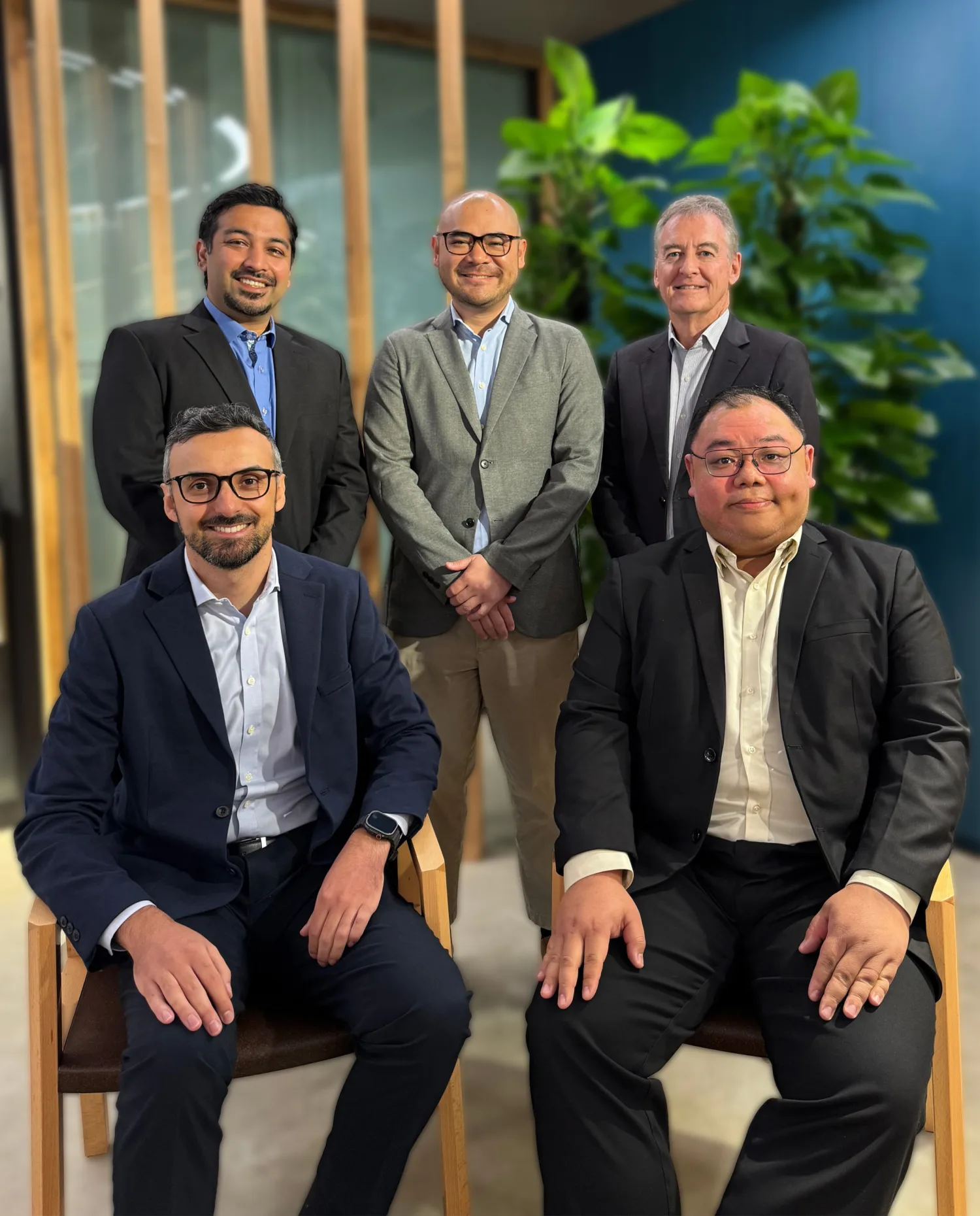 Five people in business attire pose in an office setting with two seated at the front and three standing behind them. Wooden vertical slats, overhead lighting, and tall green plants form the background.