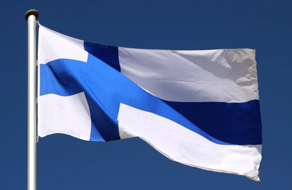 A Finnish national flag with a blue Nordic cross on a white background, mounted on a flagpole, waving against a clear blue sky.