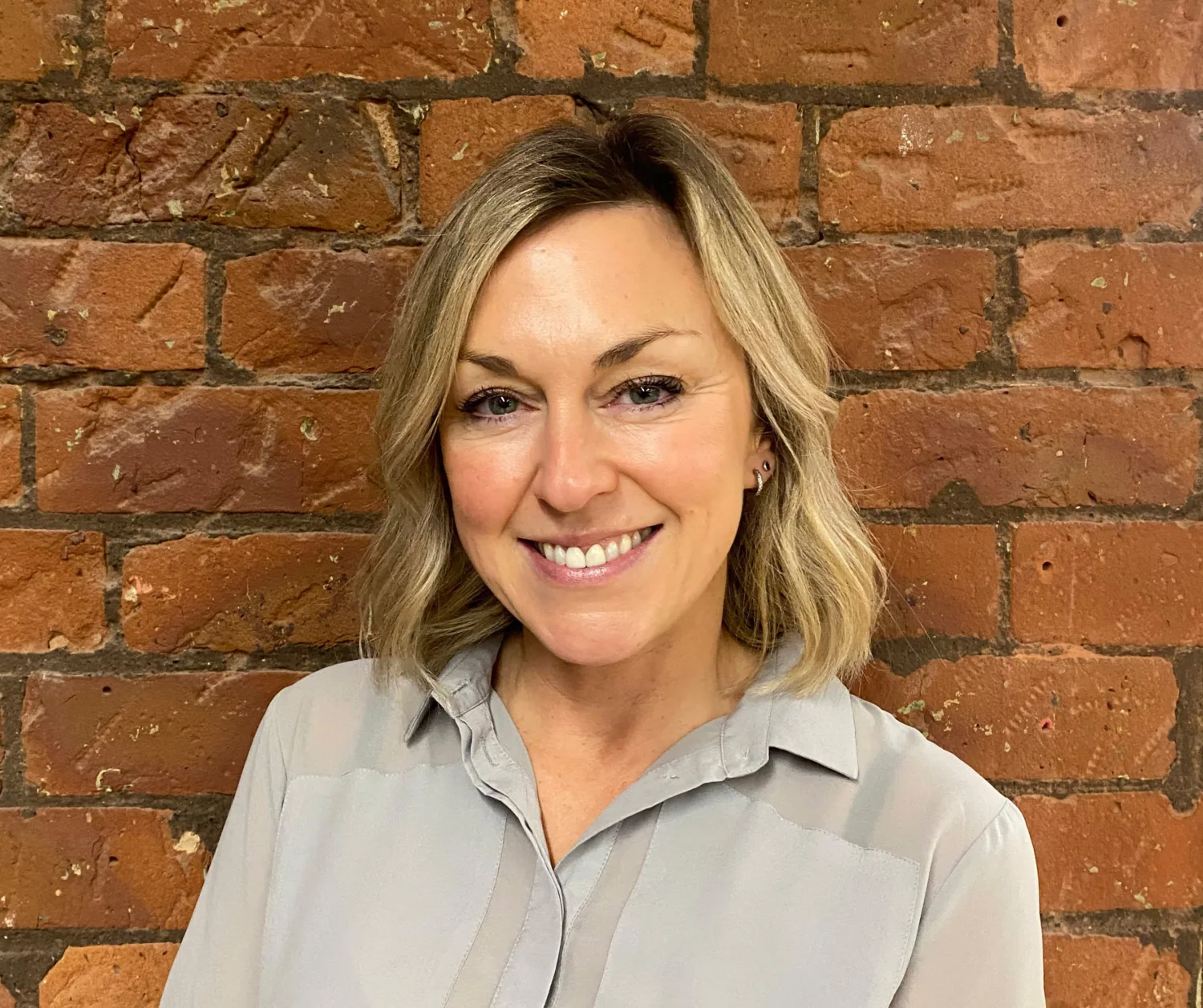 Person with shoulder-length light hair wearing a light grey collared shirt, standing against a textured red brick wall background.