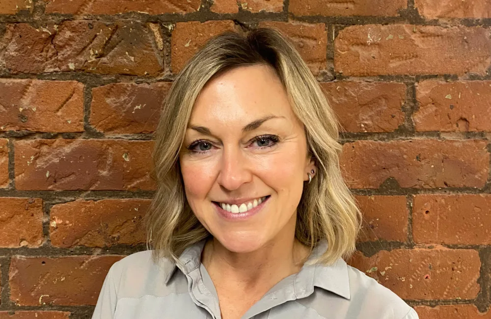 Person with shoulder-length light hair wearing a light grey collared shirt, standing against a textured red brick wall background.