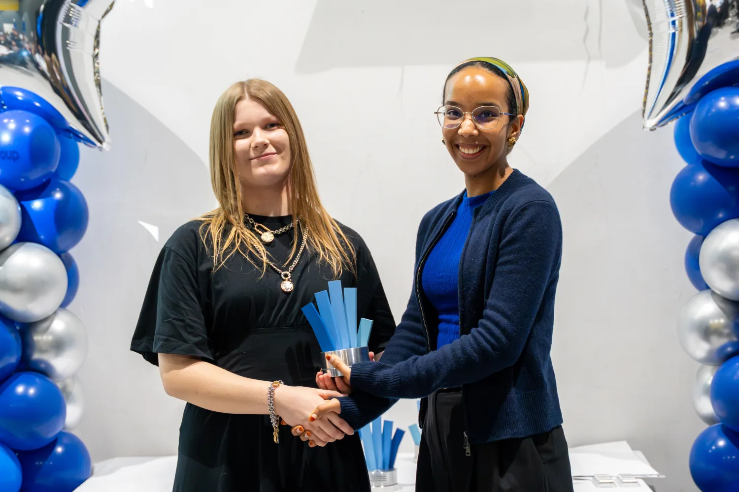 Two individuals stand side by side indoors, jointly holding a blue, fan-shaped award. They are positioned between tall balloon columns in blue, silver, and white, with a table visible in the background.