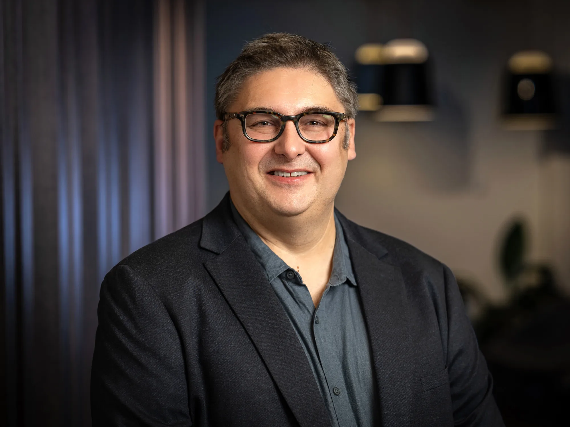 Stewart Mann wearing a dark blazer over a buttoned shirt stands indoors against a softly lit background with dark curtains and hanging light fixtures