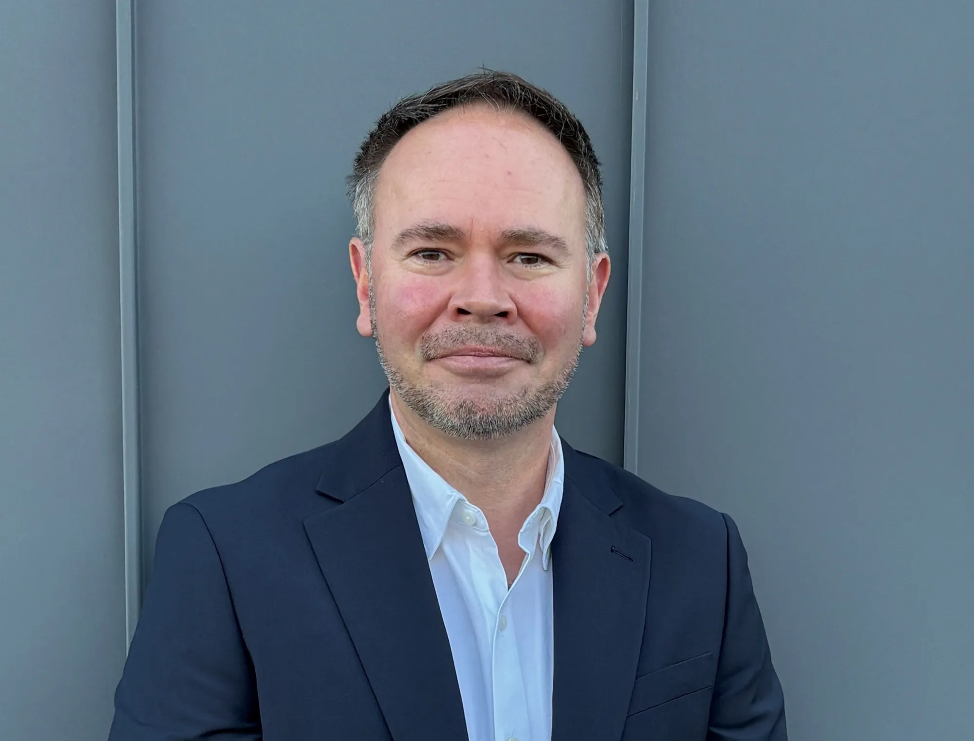 Head-and-shoulders portrait of a person wearing a navy blazer over a white collared shirt, facing the camera against a flat grey metal wall with vertical seams.