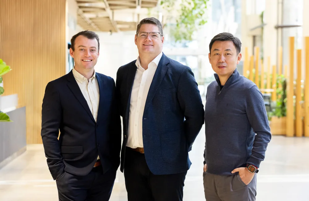 Three individuals stand indoors in a modern office space with wooden decor and plants. One wears a dark suit, another a dark blazer, and the third a casual sweater.