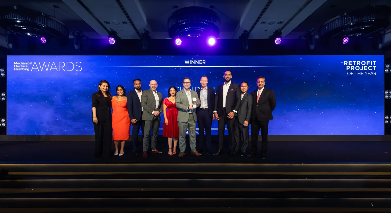 Ten people standing on a stage in front of a backdrop displaying “Mechanical Electrical Plumbing AWARDS,” “RETROFIT PROJECT OF THE YEAR,” and “WINNER”; posed for a group photo at an award ceremony.