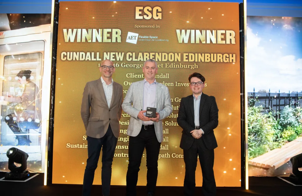 Three people stand on a stage holding an award in front of a large screen reading “ESG Winner.” The backdrop text includes “Cundall New Clarendon Edinburgh,” sponsor AET Flexible Space, and additional project details, with event lighting around the stage.