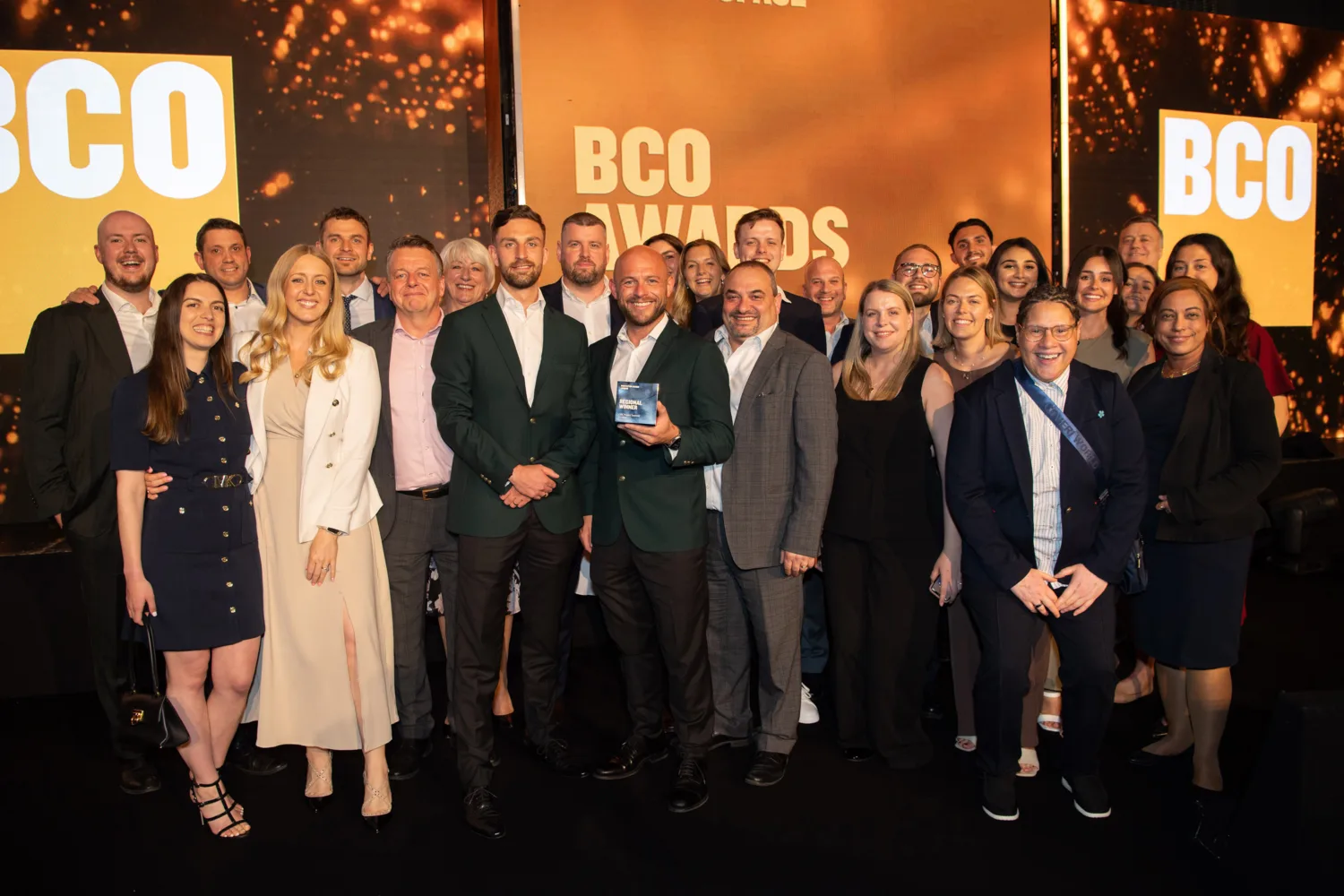 A crowd of people stood on an orange lit stage in front of BCO awards logo signage. The central person is holding the award.