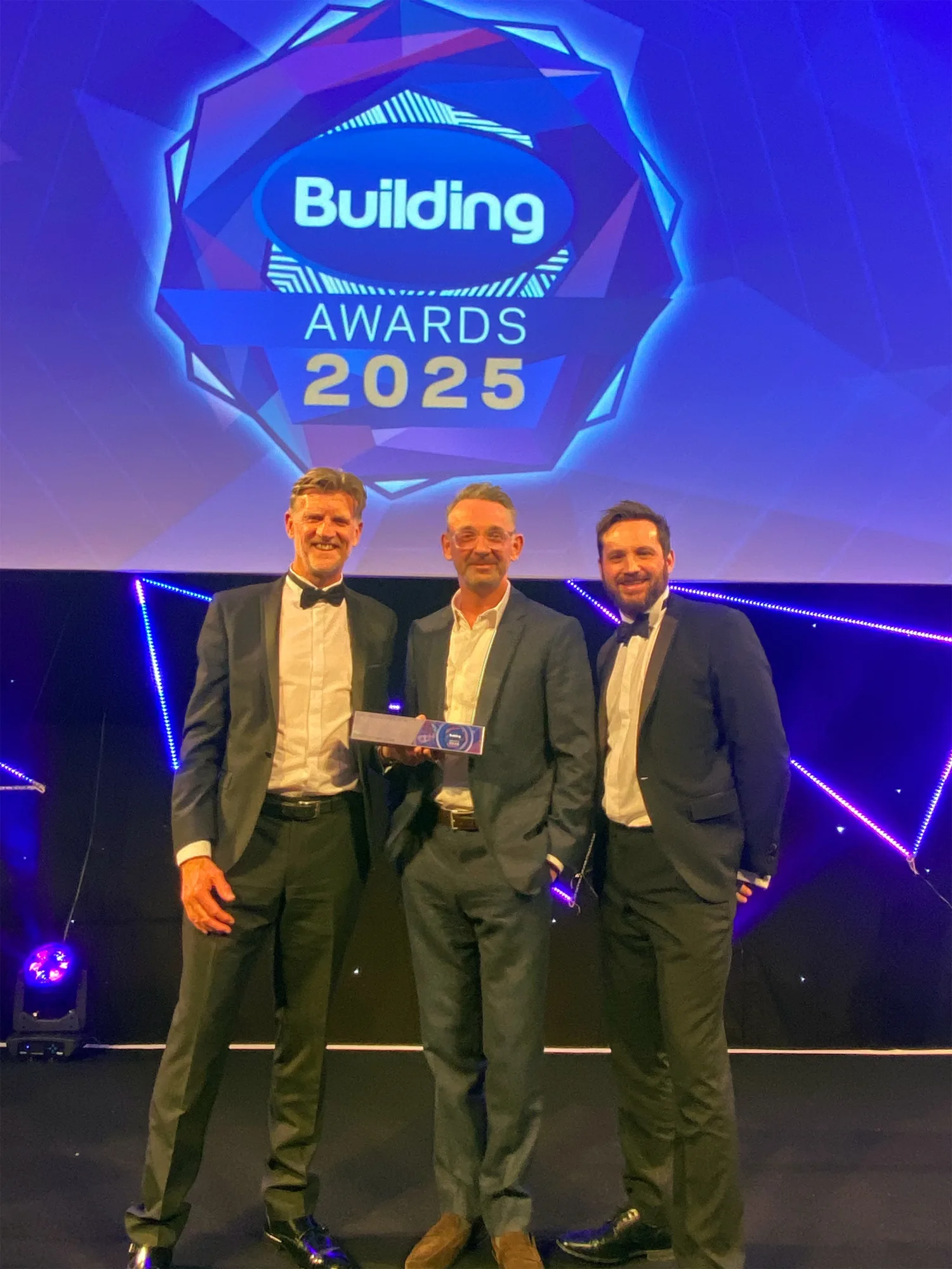 Three people in formal suits standing on a stage holding an award, with a large illuminated sign above reading ‘Building Awards 2025’ against a geometric blue and purple backdrop.
