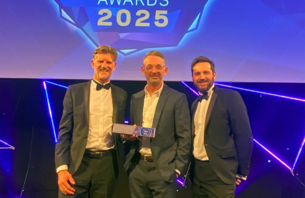Three people in formal suits standing on a stage holding an award, with a large illuminated sign above reading ‘Building Awards 2025’ against a geometric blue and purple backdrop.