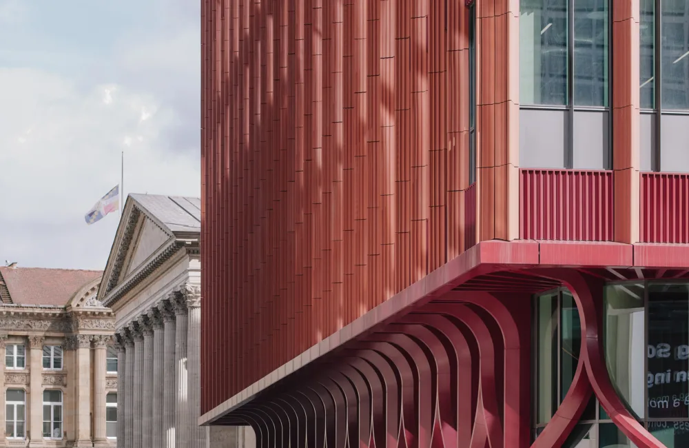 Red modern building with vertical ribbed façade and curved red supports beneath an overhang, large glass windows at right. A classical stone building with columns and a flag appears in the background under a cloudy sky.