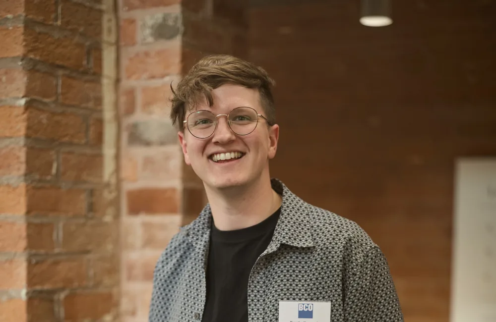 Timothy standing indoors against a brick wall, wearing a patterned short-sleeve shirt over a black t-shirt and black trousers. A name badge with text “BCO” and “Hello my name is” is pinned to the shirt.
