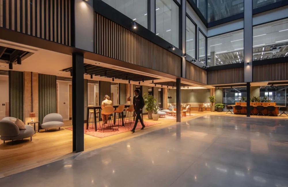 Modern open-plan office interior with double-height ceiling, glass-walled upper level, and wood slat panels. Lounge seating, tables, and plants arranged on rugs; one person seated at a table and another walking across polished floor.