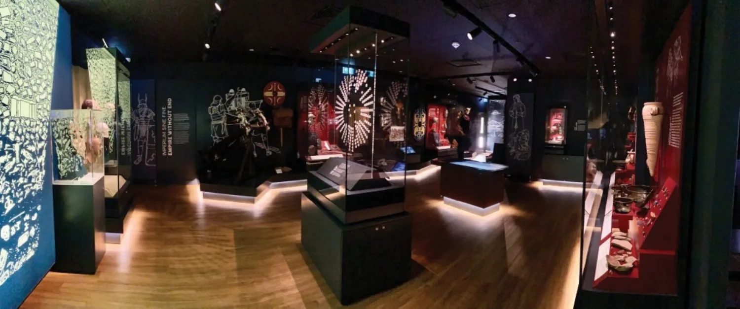 Museum gallery with wooden floor and dark walls, featuring illuminated glass display cases of artifacts. Wall panels show white illustrations of figures and objects; central case displays a circular patterned artifact.