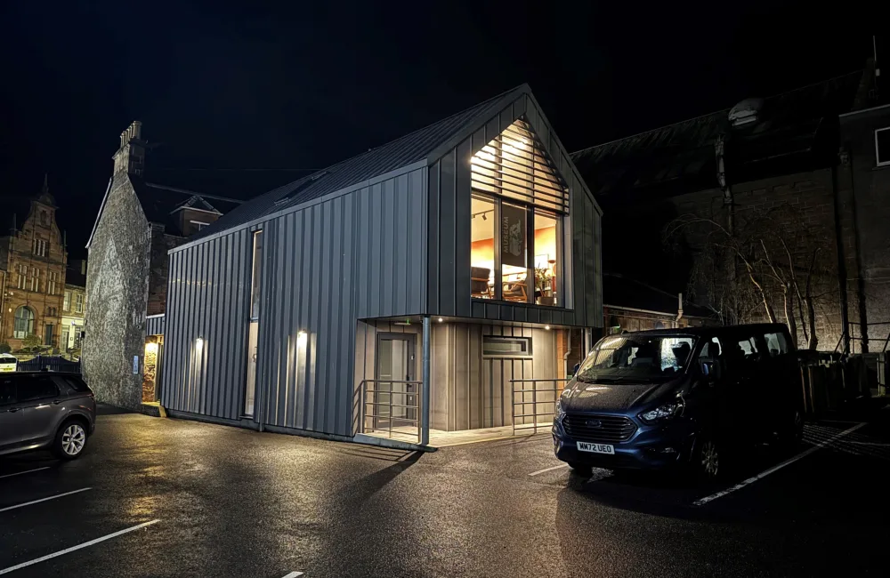 Modern two-storey building with a sloped metal roof and vertical panel siding, lit at night. Large upper window shows tables and chairs inside. Two vehicles parked on wet asphalt in front.