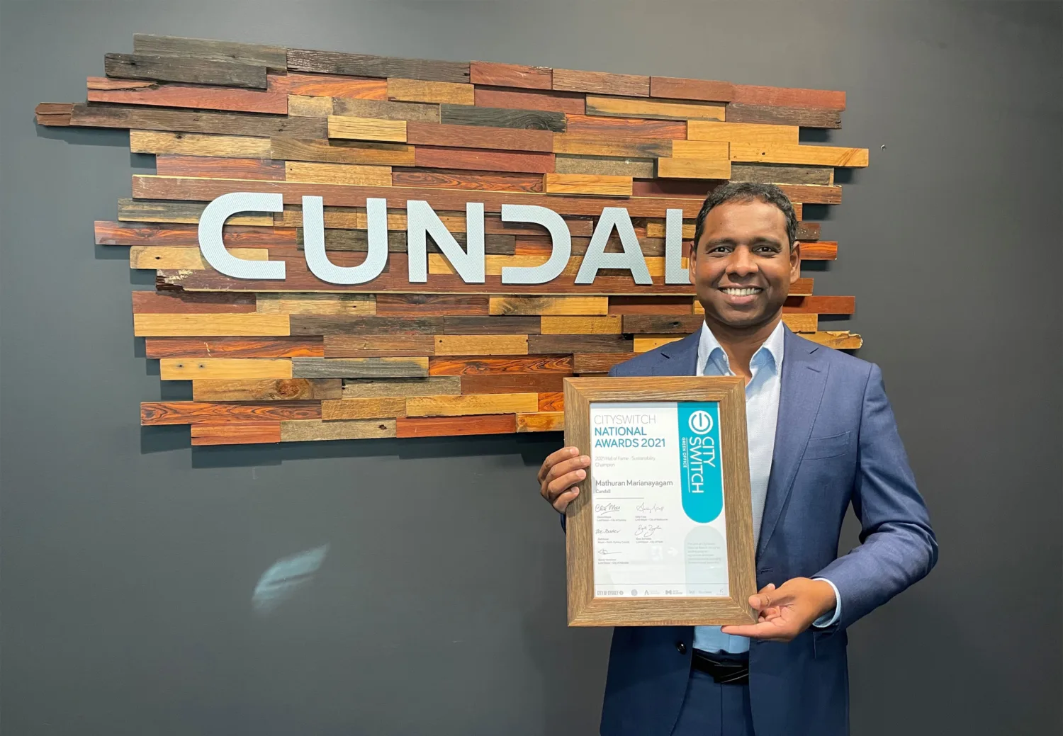 Mathuran holding his certificate in front of a wall mounted Cundall logo