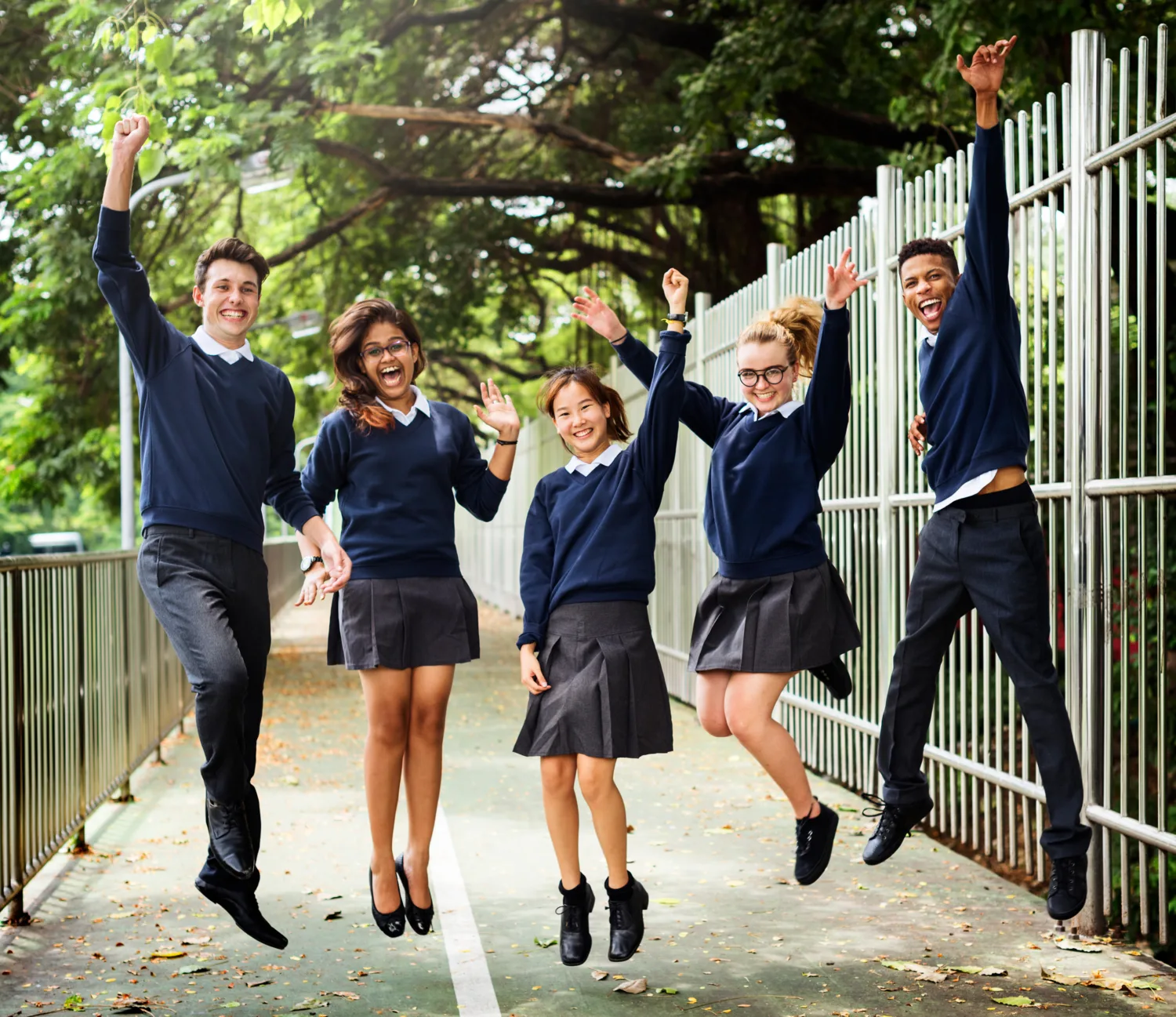 Students composed of male and female happily jumping on a walkway with trees in the background.