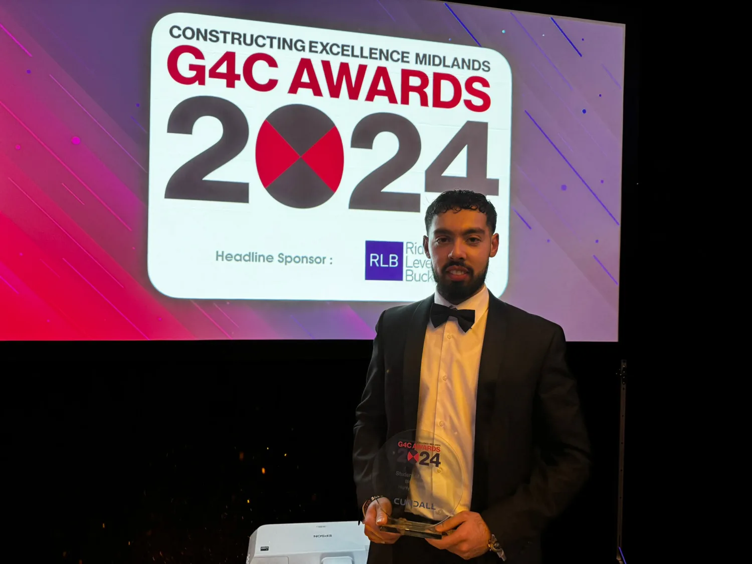 Bilal Arshad wearing a suit holding his award in front of a screen with the G4C Midlands Awards 2024 logo on it
