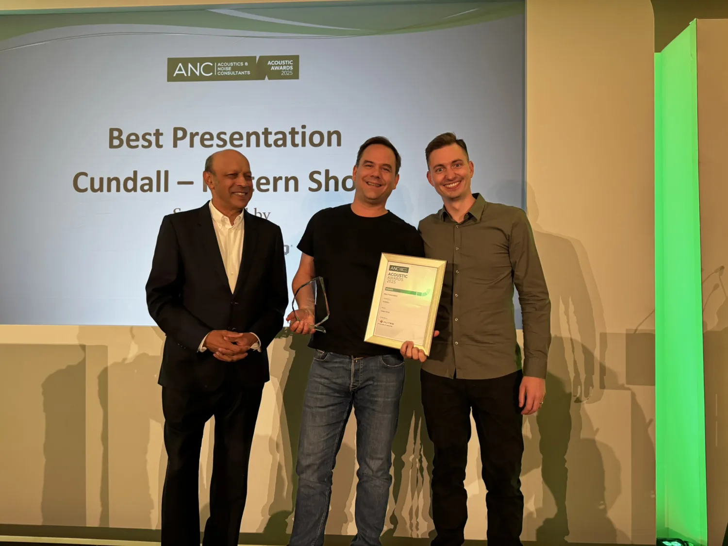 Three people stand on a stage. The person on the left wears a suit; Victor Linstrom holds a glass award and a framed certificate. A screen behind them reads: 'ANC Acoustics & Noise Consultants Association of Noise Consultants Best Presentation Cundall - Pattern Shop.