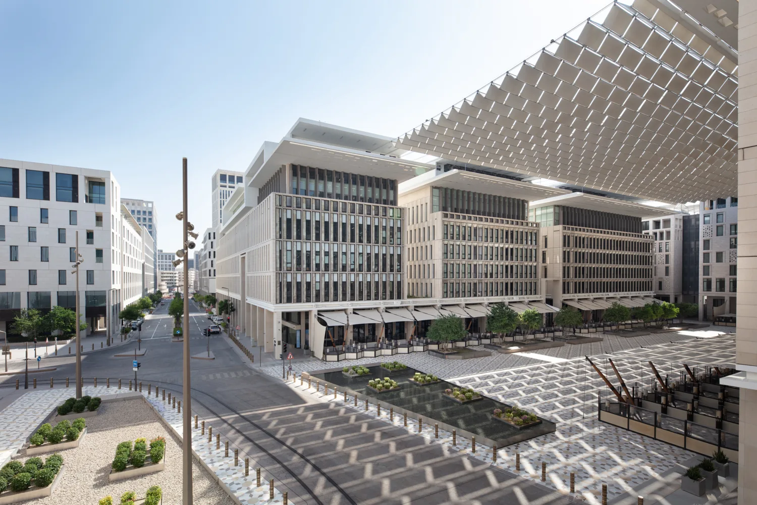 Modern urban plaza with mid-rise buildings featuring glass windows and white facades, patterned pavement, landscaped plant areas, and a large overhead shade structure.