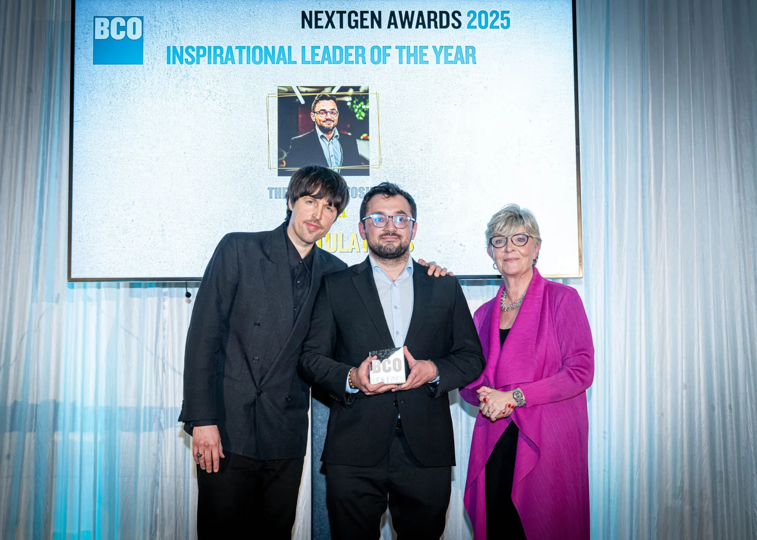 Alt text: Three people standing in front of a large screen displaying “BCO NextGen Awards 2025 Inspirational Leader of the Year” with a photo and text underneath. The person in the center is holding a rectangular award with the letters “BCO” visible. The background includes white draped curtains and bright lighting.