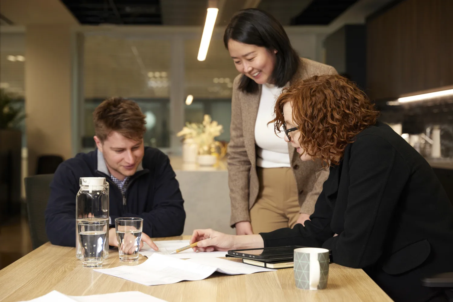 Three people gathered around a wooden table in an office setting, reviewing documents. The table holds papers, a notebook, a ceramic mug, a glass water bottle, and two drinking glasses. Background includes modern cabinetry and decorative plants.
