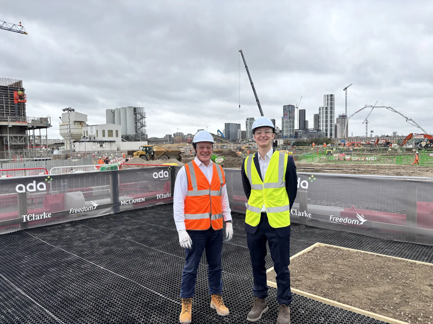 Alec Stewart stood on the left and Tom Heywood on right wearing hard hats, high‑visibility vests, long‑sleeved shirts, trousers, and work boots stand on a raised platform overlooking an active construction site with cranes, machinery, and high‑rise buildings in the background.