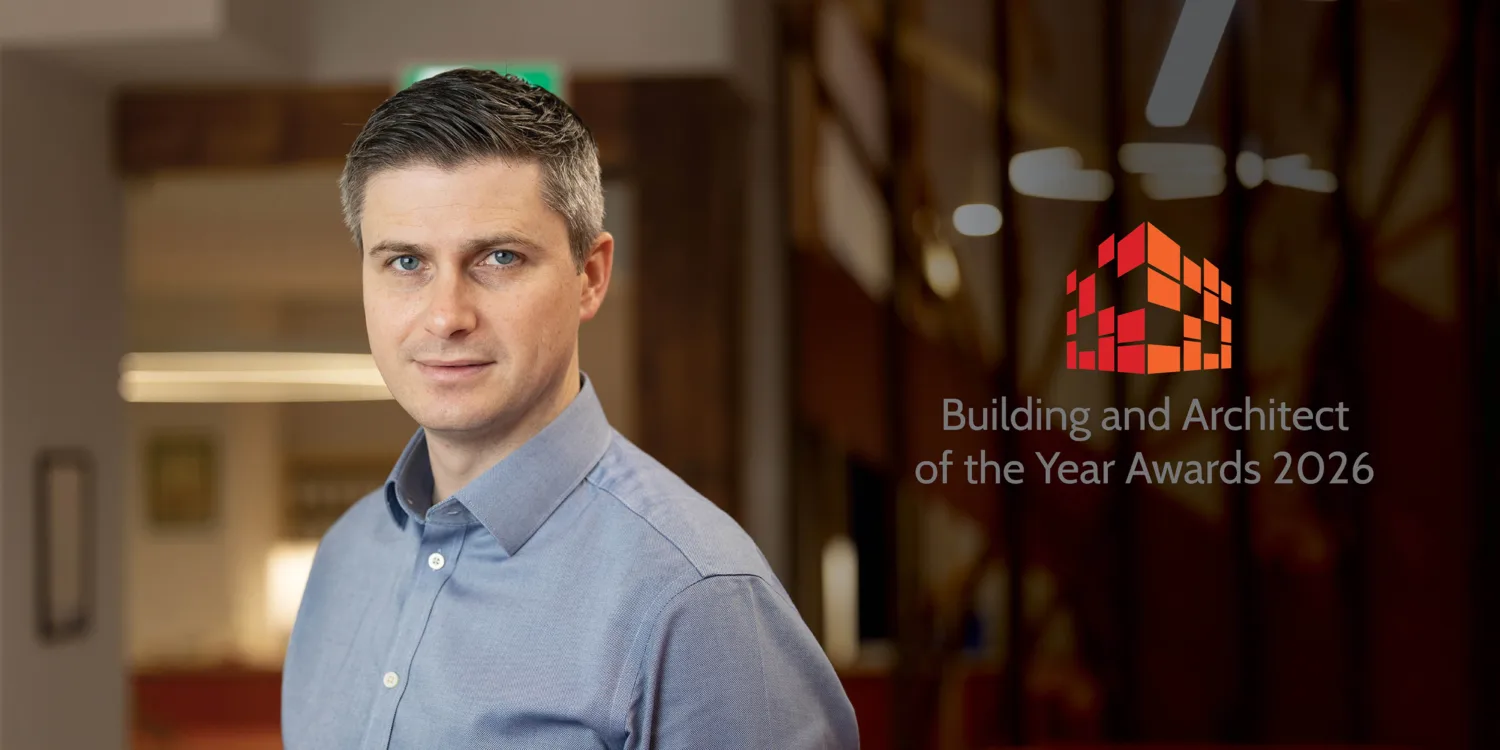 A person wearing a light blue collared shirt stands indoors beside a glass wall displaying a red geometric logo and the text “Building and Architect of the Year Awards 2026,” with warm lighting and wooden details in the background.