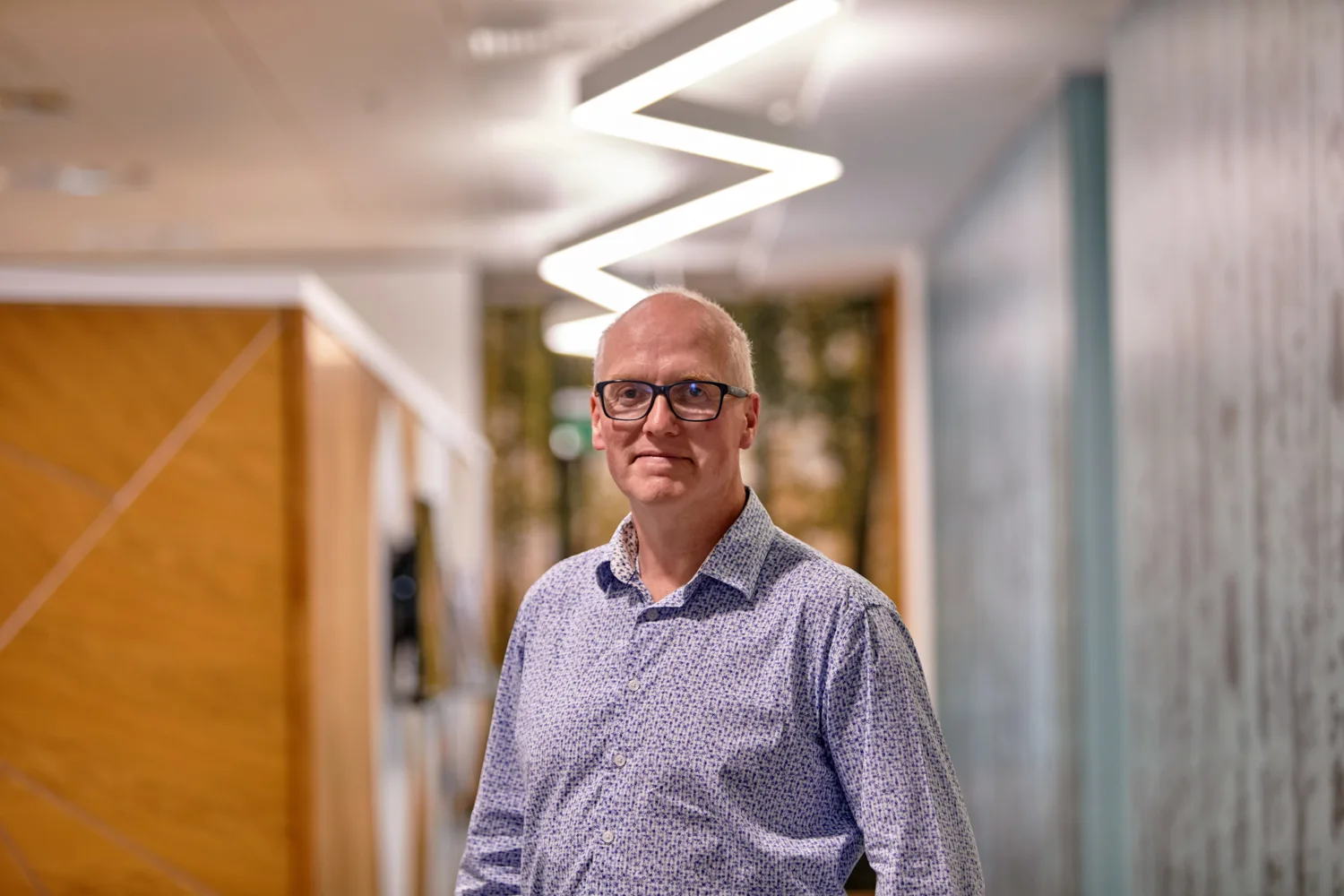 Tomas Neeson wearing a blue shirt stood in a corridor under a zigzag strip light