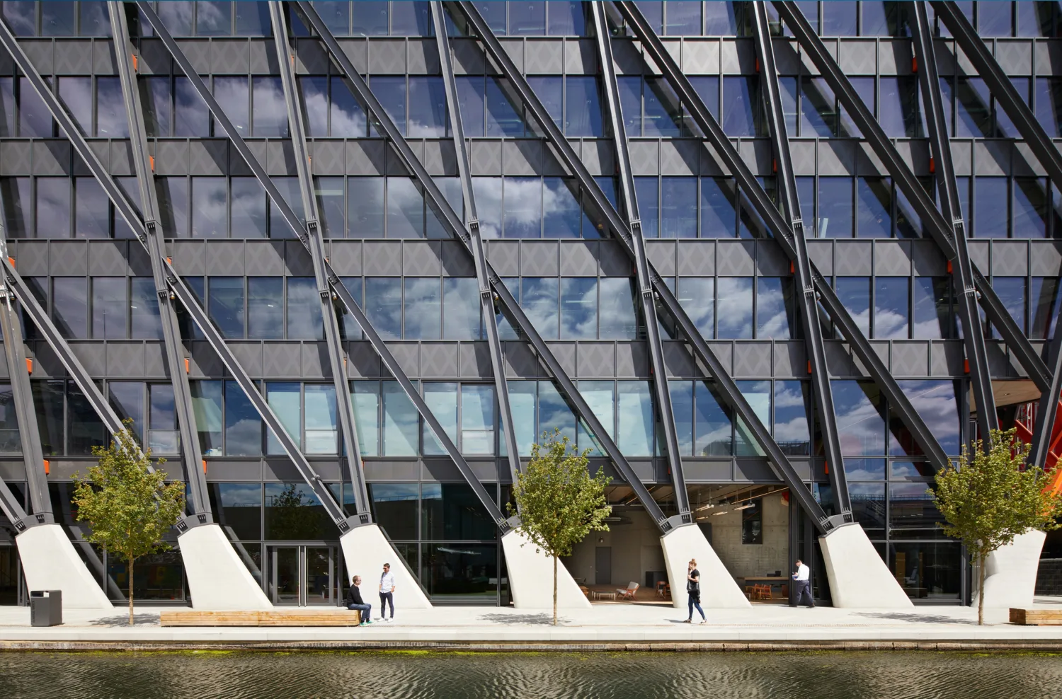 Front of the Brunel Building, a tall office building with rows of windows and metallic strips alternating vertically, while the outer building is supported by metallic beams in a geometric pattern. At the base of the photo is a body of water and a white paved path.